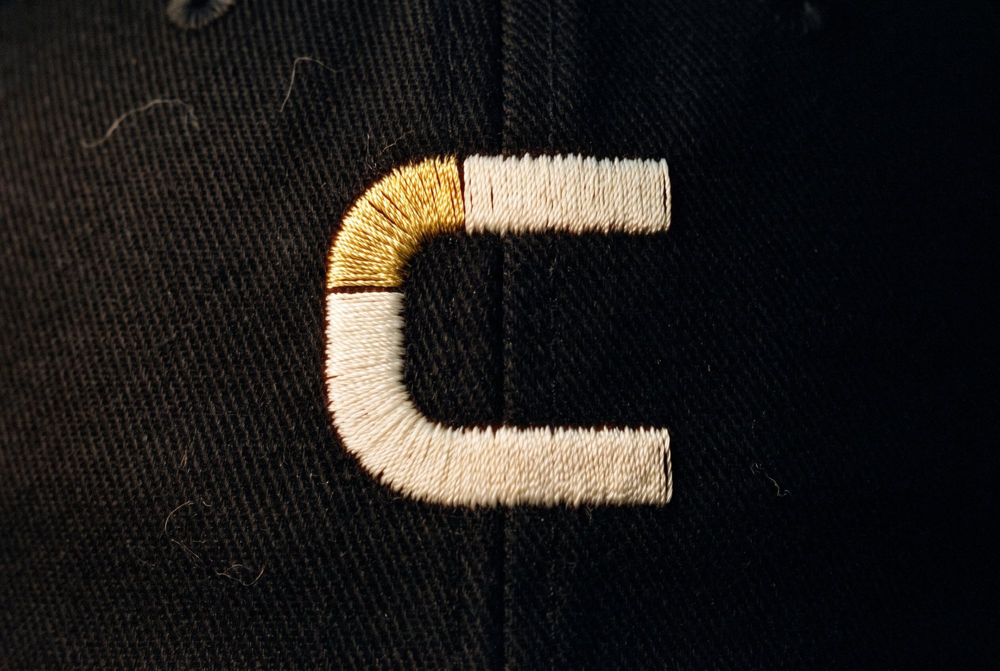 The Coldwork mark embroidered in bone satin stitch with a gold thread bend on black twill.