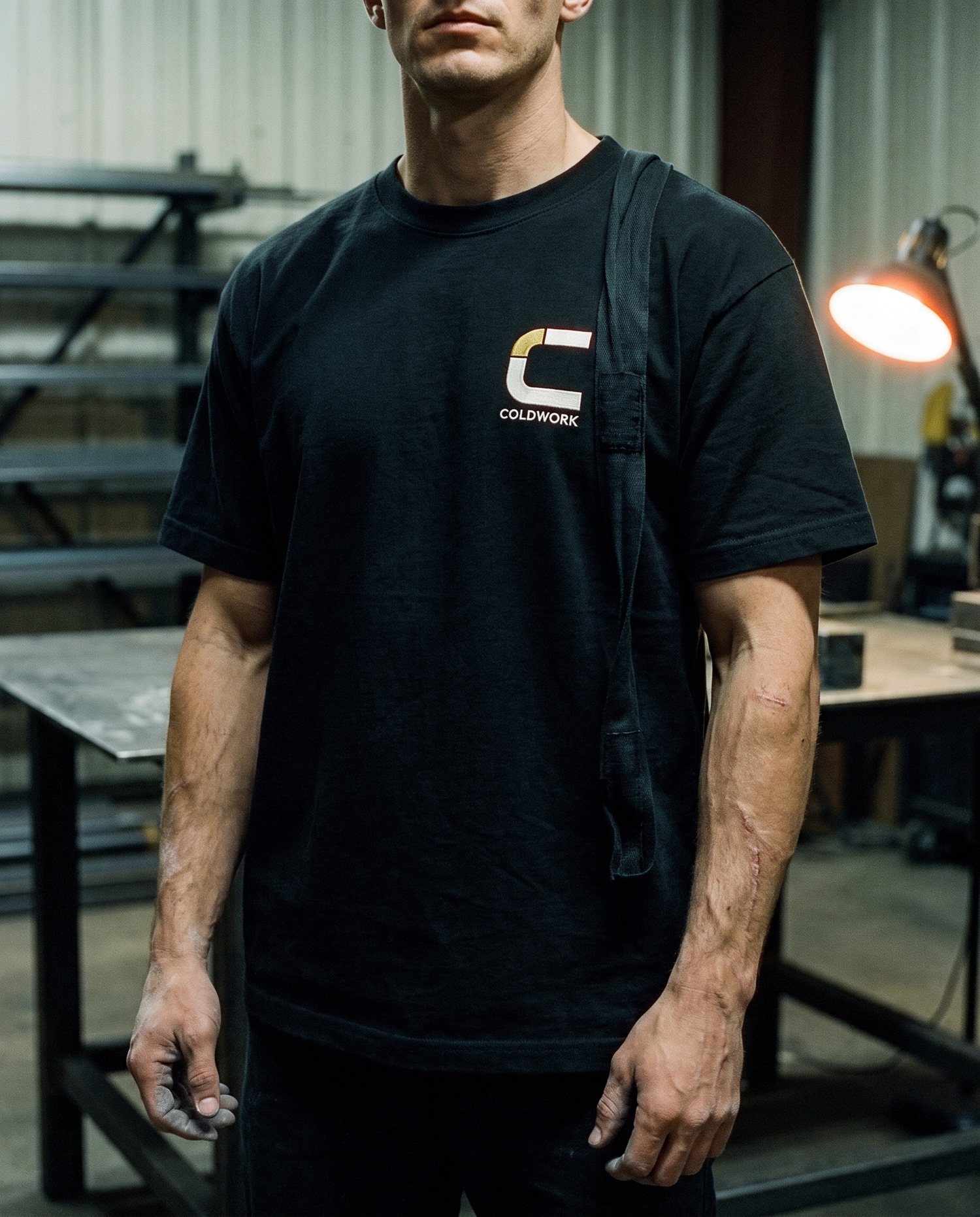 A black heavyweight tee with the Coldwork mark embroidered on the left chest, worn in a welding shop with hand wraps around the neck.